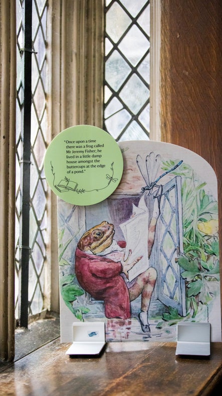 A colourful illustration of a frog sitting reading a newspaper by an open window, in front of a leaded window